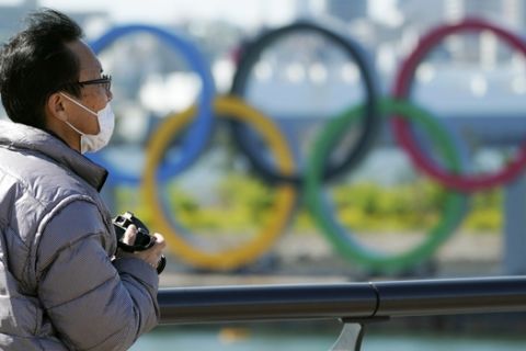 A visitor wearing a face mask stands near the Olympic rings at Tokyo's Odaiba district Tuesday, March 24, 2020. The Tokyo Olympics are probably going to happen, but almost surely in 2021 rather than in four months as planned. (AP Photo/Eugene Hoshiko)