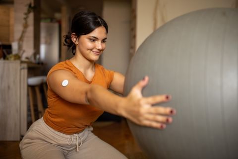 Young diabetic patient teenage woman exercising with fitness ball at home, she has a glucose monitor on her arm.