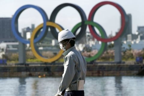 A masked man works at a construction site with the Olympic rings in the background Tuesday, March 3, 2020, at Tokyo's Odaiba district in Tokyo. The spreading virus from China has put the Tokyo Olympics at risk. The Olympics are to open on July 24 - less than five months away. (AP Photo/Eugene Hoshiko)
