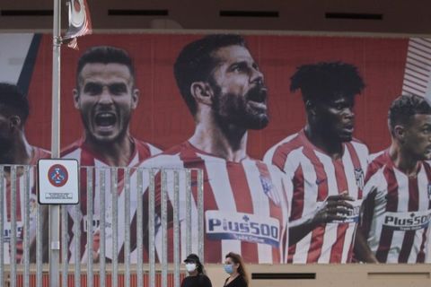 Two women wearing face masks pass by a giant poster of Atletico Madrid soccer players at the Wanda Metropolitano stadium in Madrid, Spain, Tuesday, May 5, 2020. The Spanish soccer league aims to restart in June without spectators. It's new compulsory protocols say all players, coaches and club employees must be tested for COVID-19 before training resumes, then regularly after that. All clubs' training facilities must be properly prepared and disinfected before players can start practising individually. (AP Photo/Paul White)