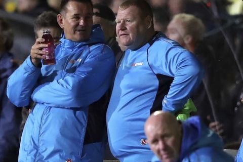 Sutton reserve goalkeeper Wayne Shaw, top right, and manager Paul Doswell, left, stand near the team dugout during the FA Cup 5th round soccer match at Gander Green Lane, Sutton, London, Monday Feb. 20, 2017. A  backup goalkeeper paid the price for eating a meat pie during his team's landmark game against Arsenal, resigning from Sutton United as authorities launched investigations into the apparent betting stunt. A British newspaper's betting company, which sponsored Sutton United for Monday's FA Cup game, had 8-1 odds that overweight reserve goalkeeper Wayne Shaw would eat a meat pie. (Andrew Matthews/PA via AP)