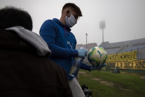 A ball catcher wearing plastic gloves and a face mask amid the new coronavirus pandemic, disinfects the ball during a local championship soccer match between Nacional and Penarol in Montevideo, Uruguay, Sunday, Aug. 9, 2020. (AP Photo/Matilde Campodonico)