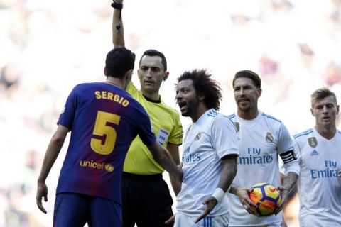 Real Madrid's Marcelo , center, gestures while Referee Jose Maria Sanchez Martinez shows a yellow card to Barcelona's Sergio Busquets, left, during the Spanish La Liga soccer match between Real Madrid and Barcelona at the Santiago Bernabeu stadium in Madrid, Spain, Saturday, Dec. 23, 2017. (AP Photo/Francisco Seco)